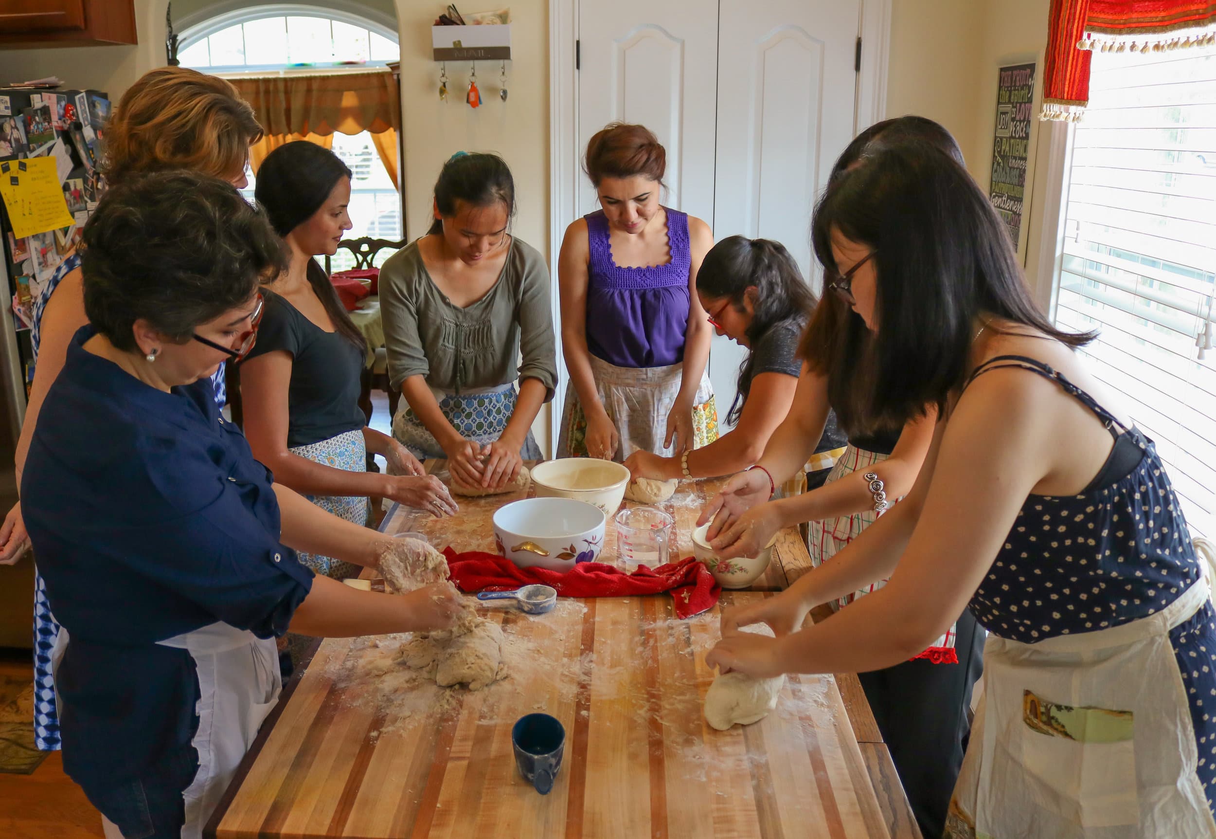 Women in aprons knead bread dough around a long wooden kitchen table dusted with flour.